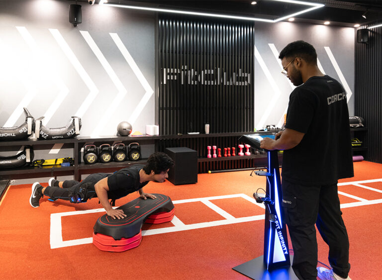 “A fitness trainer supervises a person performing push-ups on a step platform during an EMS training session at FitClub, Gurgaon.”