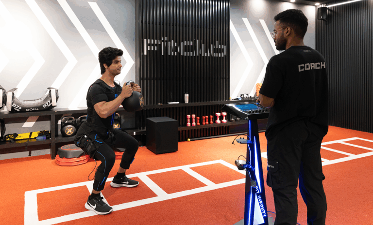 “Man performing squats with a kettlebell while wearing EMS training suit under the supervision of a fitness coach in a gym.”