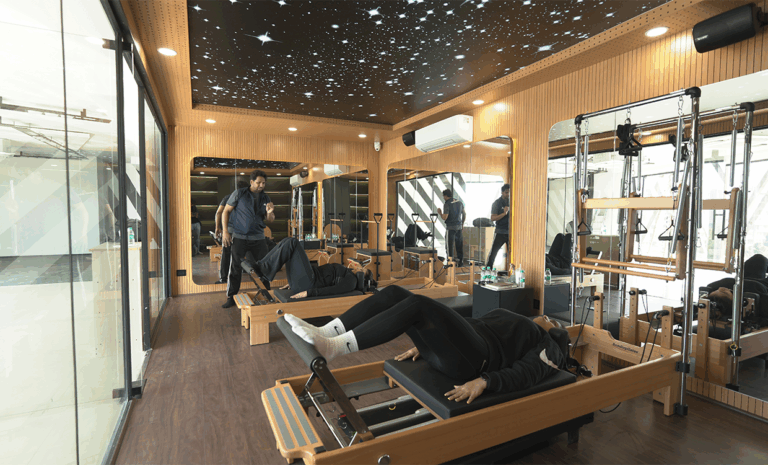 “People exercising on Pilates reformer machines in a modern fitness studio with mirrored walls and starry ceiling lights.”
