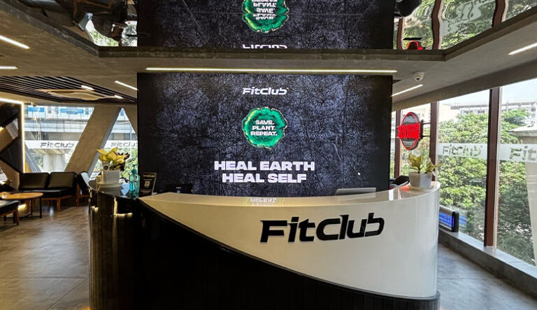 “FitClub reception desk with a digital display showing the message ‘Heal Earth Heal Self’ and the slogan ‘Save Plant Repeat.’”
